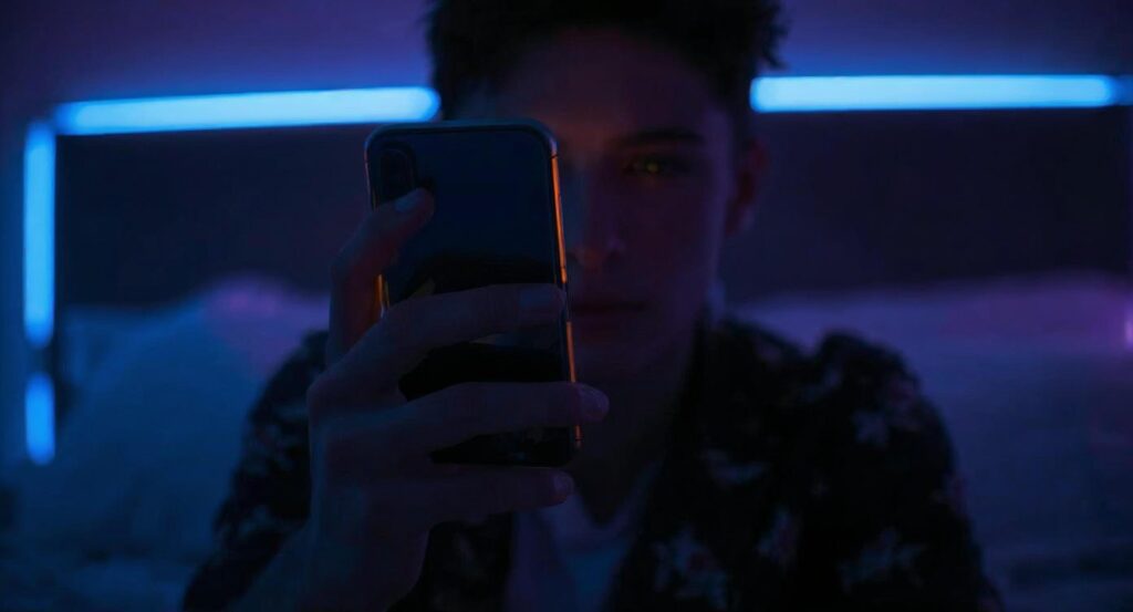 Gros plan sur la main d'un jeune Français de 18 ans, tenant délicatement un smartphone lumineux. En arrière-plan, une chambre moderne à l'éclairage tamisé.